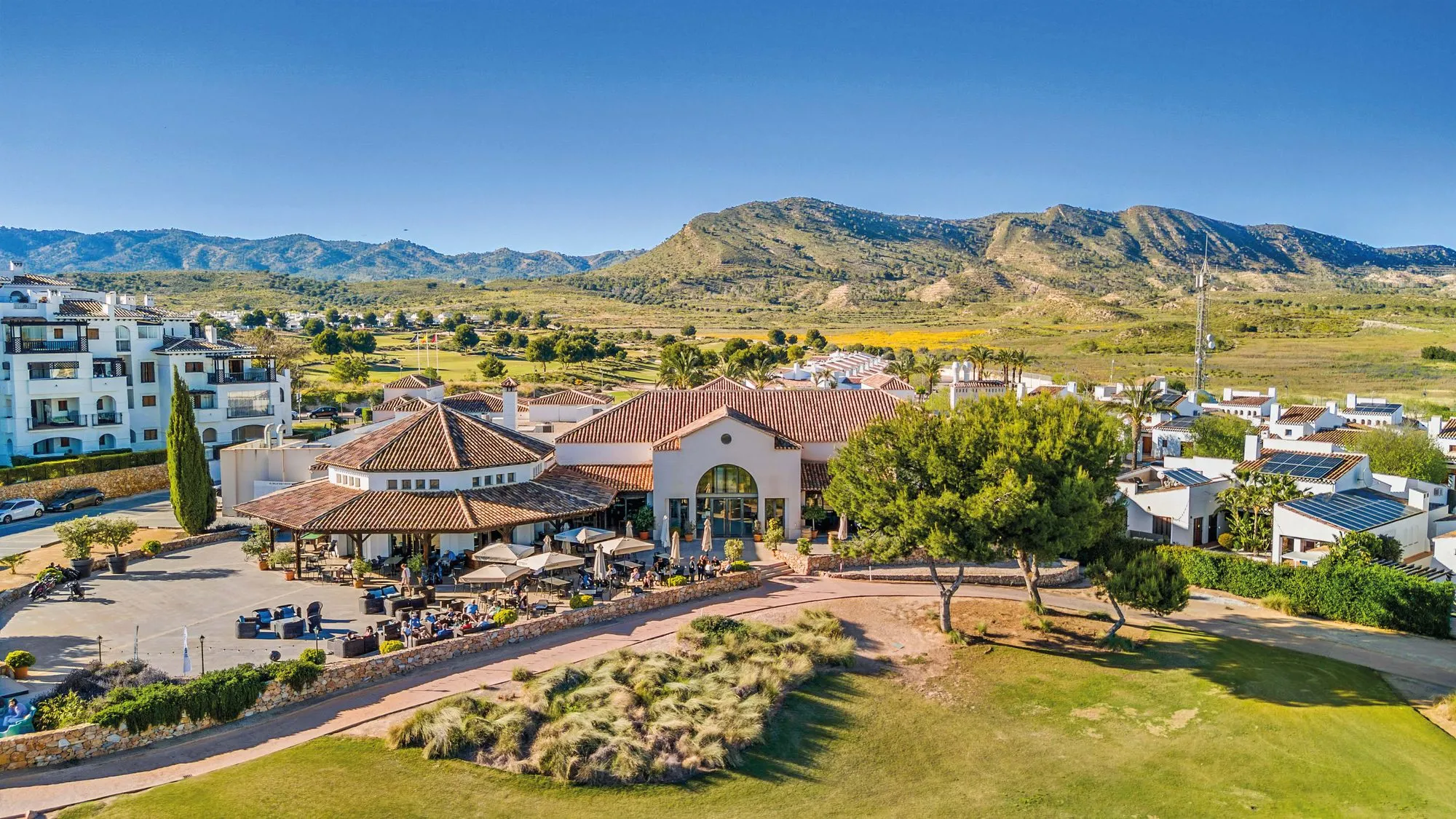 Aerial photo of the El Valle Golf Resort clubhouse, with tiled roofs, a terrace and car park, and mountains behind