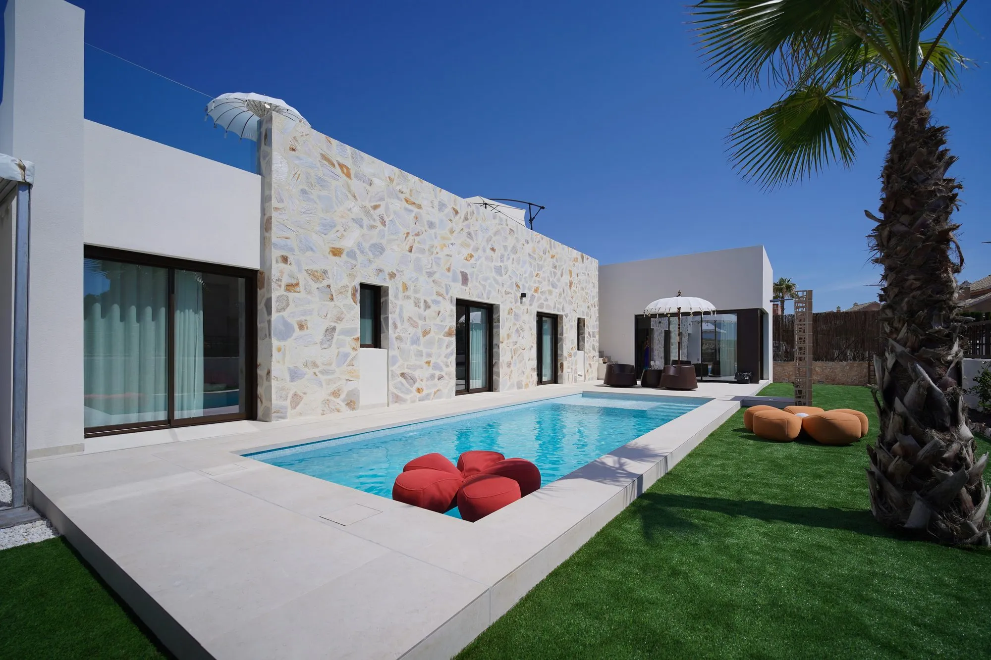 The LIEGE Mediterránea show home: the private pool beside the natural stone façade, with lawn, a palm tree and a parasol
