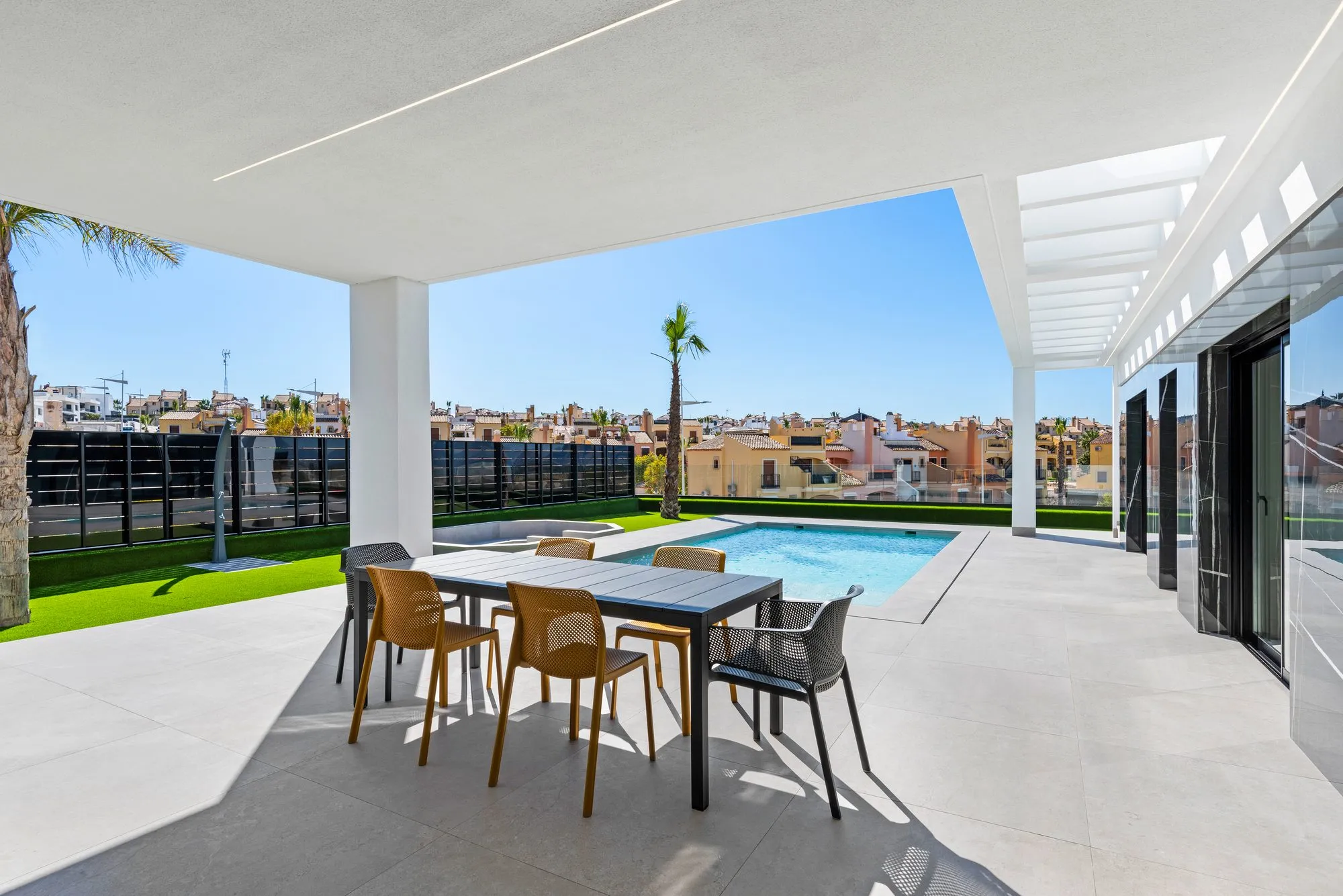 Covered porch with an outdoor dining table and yellow and black chairs, facing the pool and garden