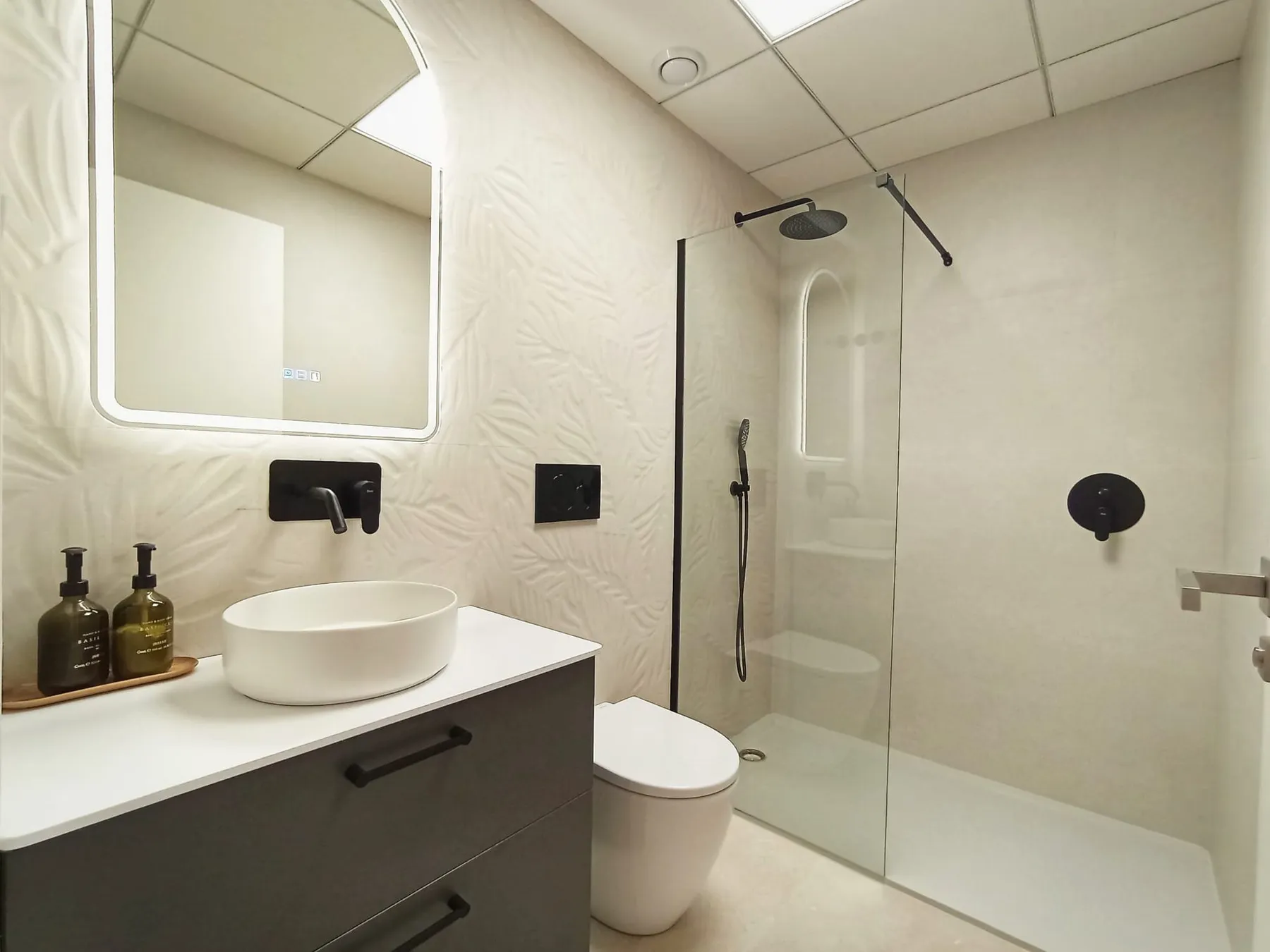 Photo of the show flat bathroom, a vessel basin on a dark vanity unit, a backlit mirror, textured white tiles and a walk-in shower with black fittings