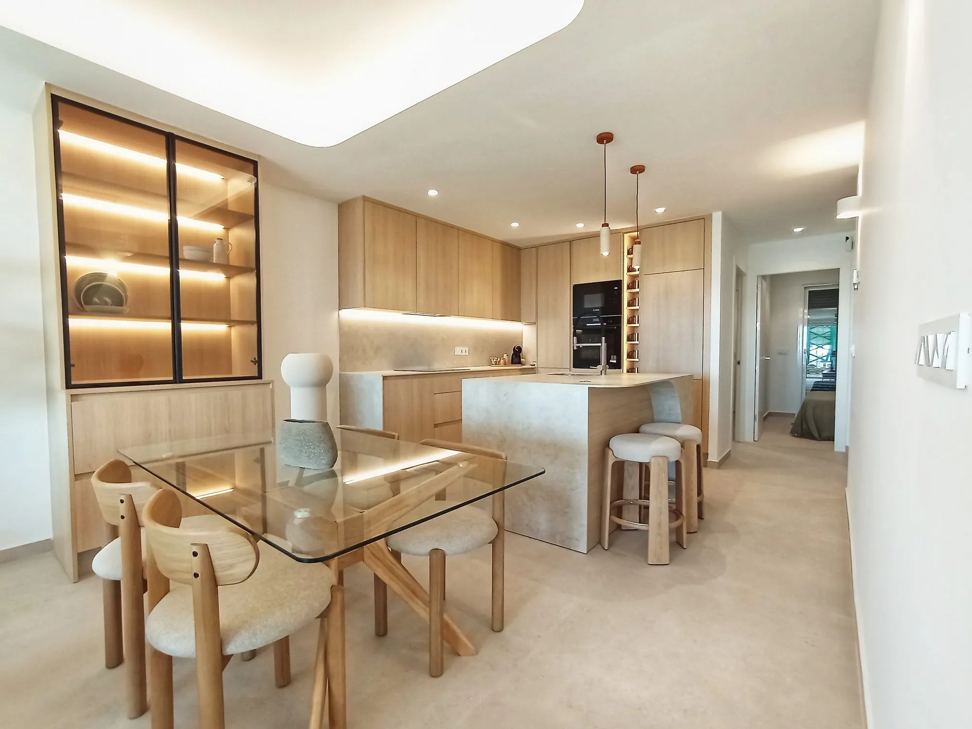 Photo of the show flat dining area, a glass table with wooden chairs, a lit glass-fronted cabinet and the open kitchen with an island behind