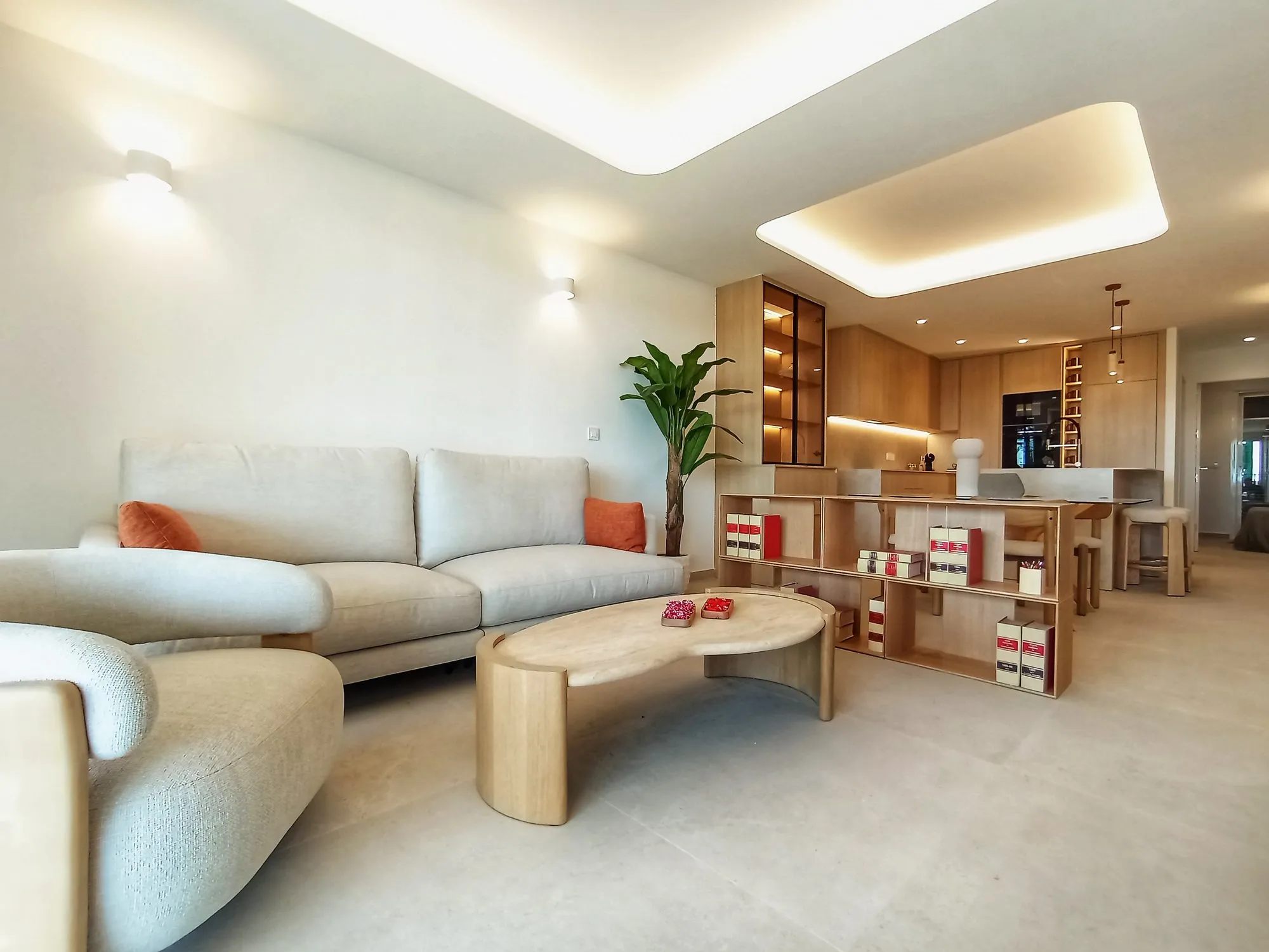 Photo of the show flat living room, a light sofa and curved coffee table, a wooden shelf unit and the open kitchen behind, under a lit ceiling recess