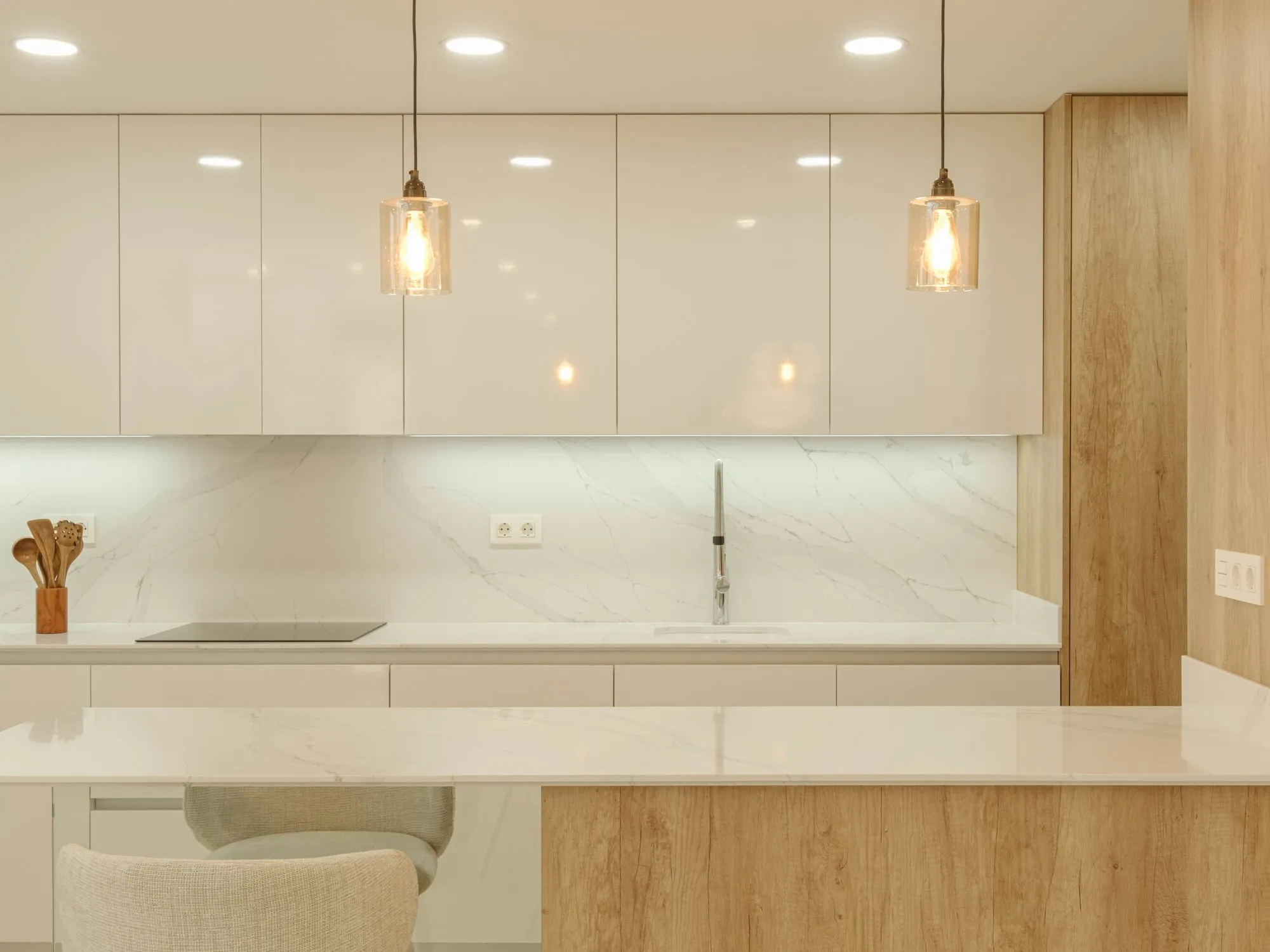Photo of the show house kitchen, gloss white units, a wood-finish tall unit, a white quartz worktop and splashback, and pendant lights