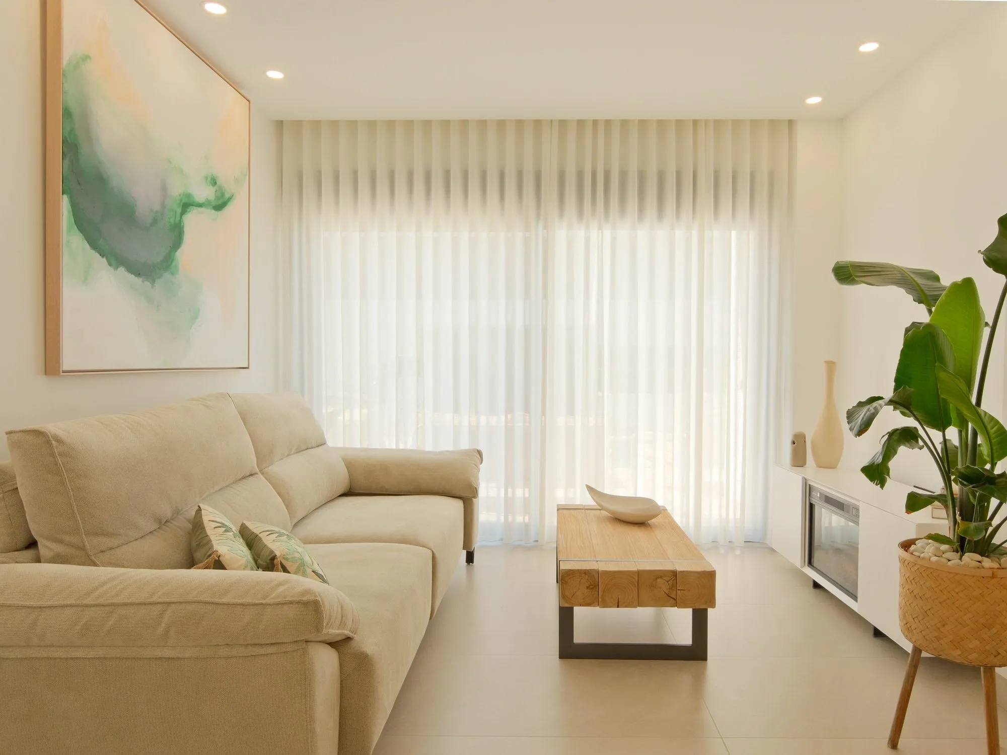 Photo of the show house living room, a beige sofa, a wooden coffee table, a white TV unit and light curtains over the large window