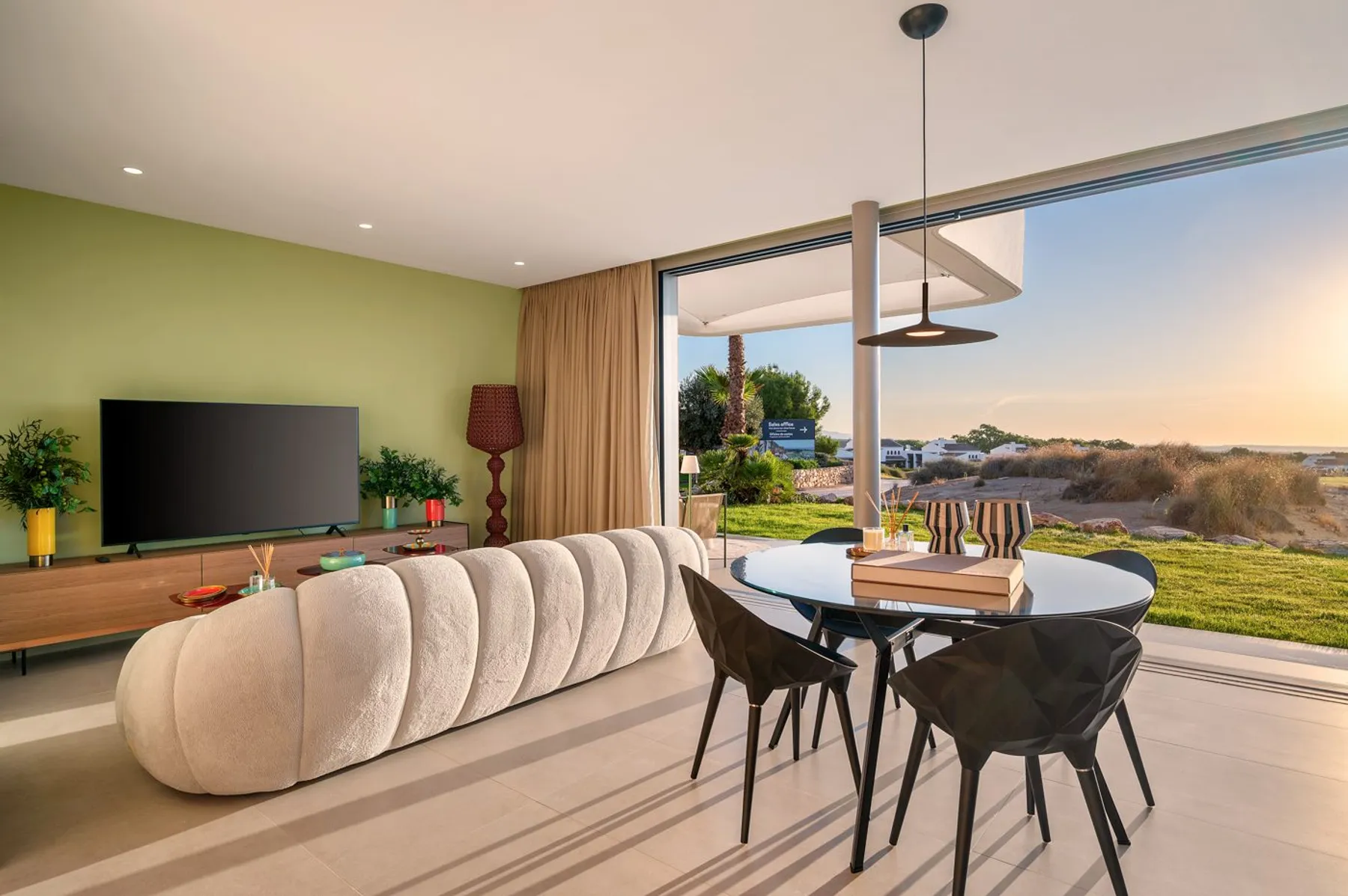 Photo of the show villa’s living and dining room, with a curved cream sofa, a lime-green wall, a dark round table and large glass doors to the garden