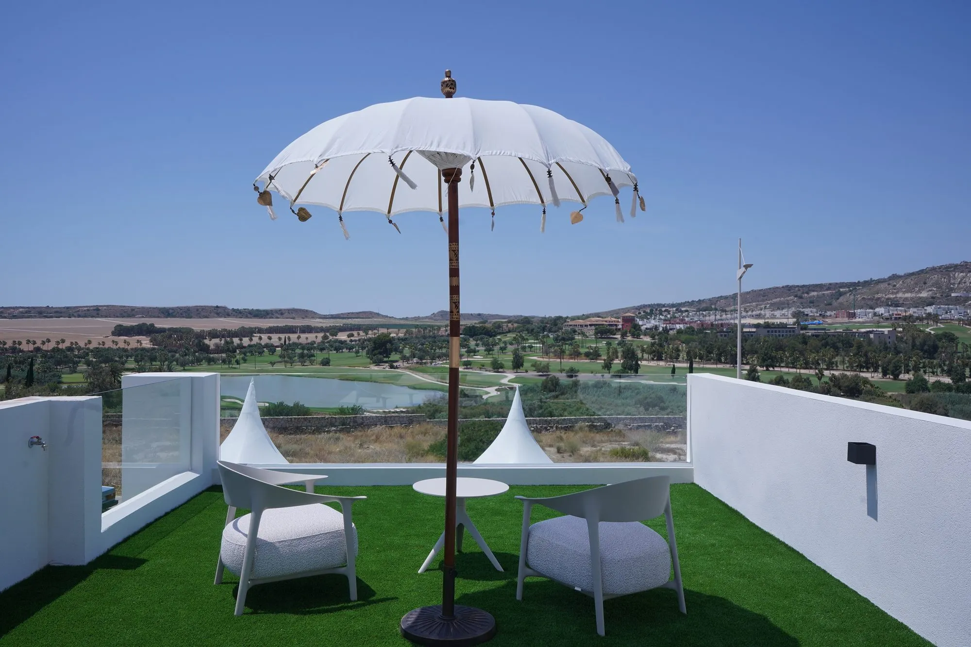 The rooftop solarium of the show home, with artificial grass, two armchairs, a parasol and a glass railing, and the golf course and lake behind