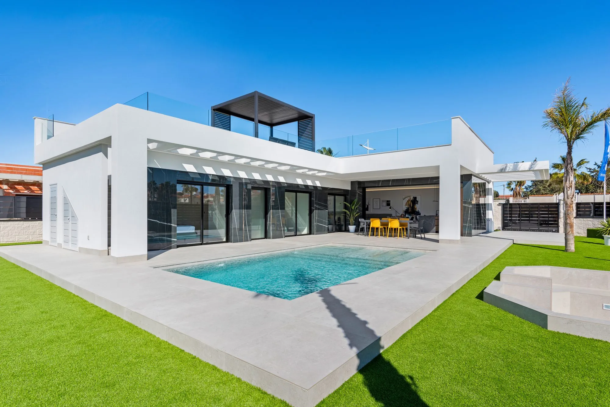 White single-storey villa with a glazed, black marble-effect façade, a rectangular pool, stoneware terrace, lawn and a pergola on the roof